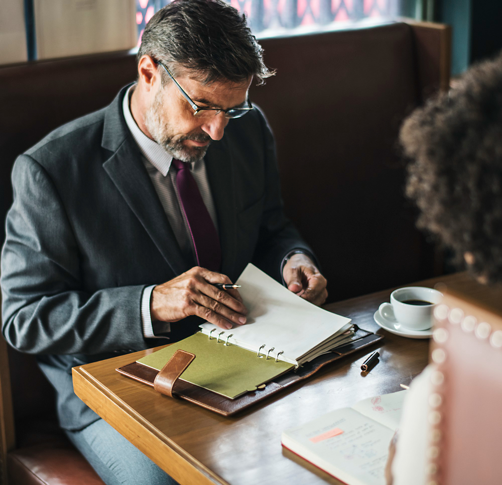 A man in formal attire and a woman at a table, engaged in conversation about notarization and document services.